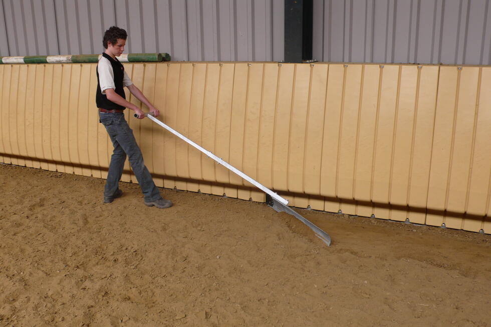 Ein metallischer Hufschlagboy mit einer abgeschrägten Schaufel, einem Metallstiel und einem rechtwinklig darauf montierten Griff, jeweils am Ende schwarz gummiert, wird von einer Person der Kante einer Reithalle nach, durch den Sand gezogen