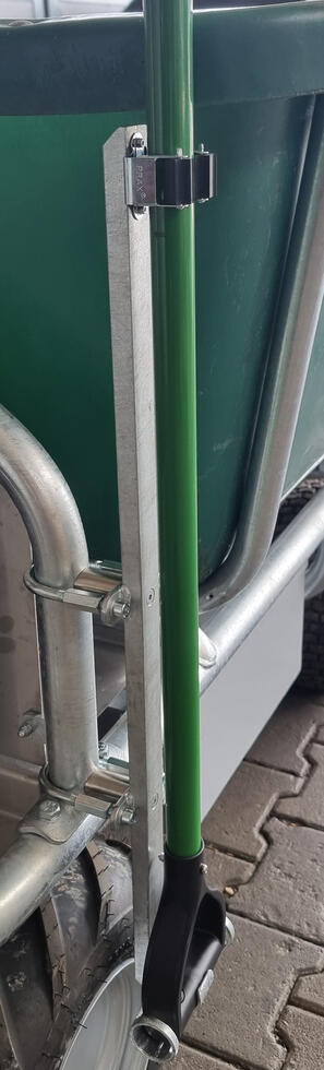 Eine grüne Kunststoffschubkarre mit vier Rädern und einem Trittbrett, hinter der Mulde befindet sich die Steuerungseinheit auf einem Vierkant mit schwarzen Griffen und orangem Halter. Das Gestell ist aus Metall, die Reifen schwarz. Eine Person fährt auf einem Trittbrett stehend eine Rampe an einem Miststock hoch