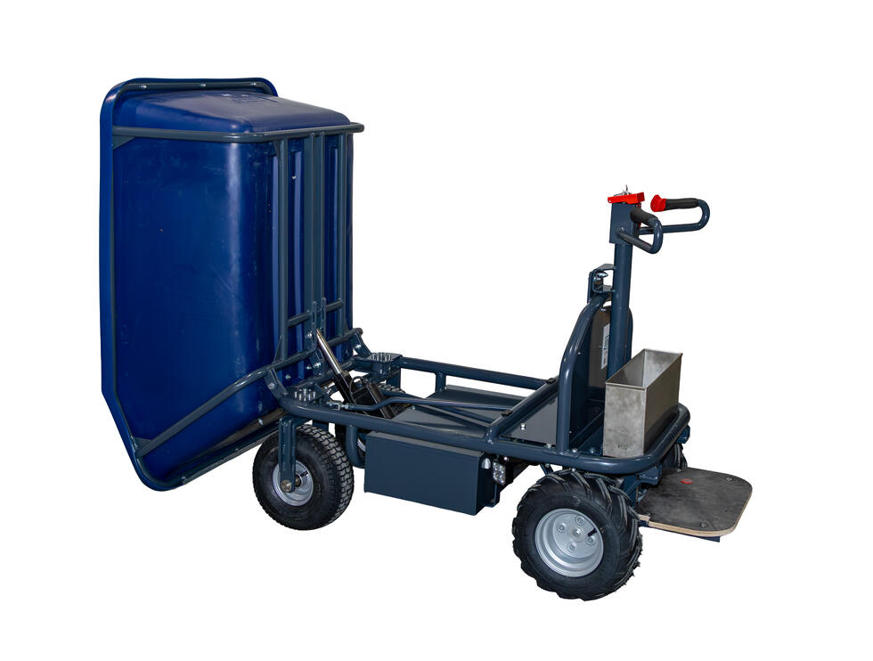 Eine grüne Kunststoffschubkarre mit vier Rädern und einem Trittbrett, hinter der Mulde befindet sich die Steuerungseinheit auf einem Vierkant mit schwarzen Griffen und orangem Halter. Das Gestell ist aus Metall, die Reifen schwarz. Eine Person fährt auf einem Trittbrett stehend eine Rampe an einem Miststock hoch