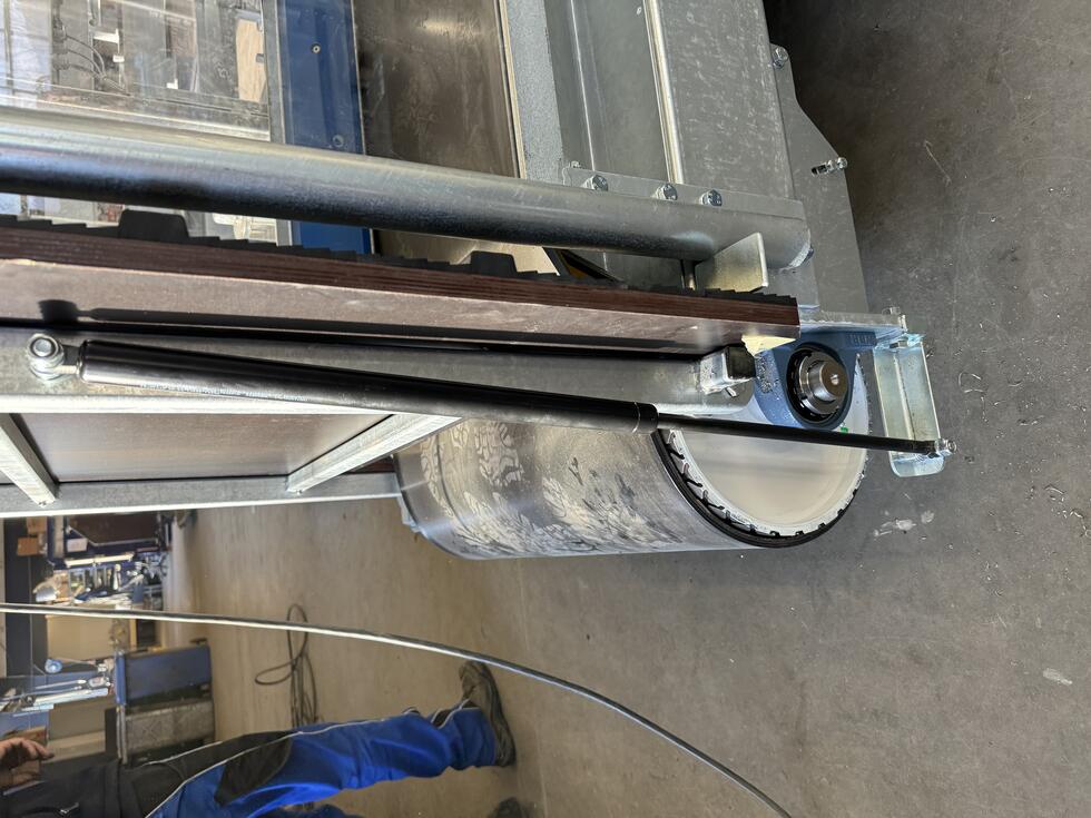 Ein Pferdesolarium, zweifarbig, grüner und grauer Kunststoff, leicht gewölbt und gebogen, mit roten und andersfarbigen Lampen, sowie Ventilatoren auf der Unterseite, aufgehängt in Stallgang, roter Boden, rechts und links Pferdeboxen, auf jeder Seite schaut jeweils ein schwarzes Pferd in den Gang hinaus