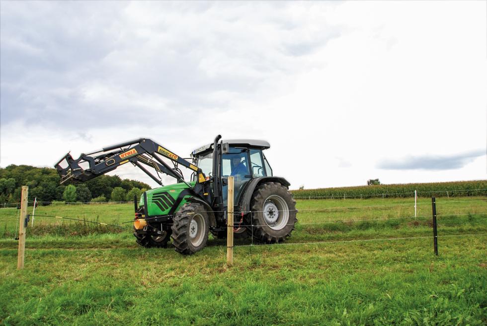 Ein grüner Traktor der einen Zaun durch das zweiteilige, an zwei Pfähle montierte Weidetor passiert, Mais und Wald im Hintergrund