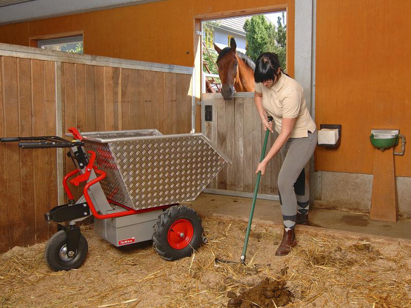 Eine Schubkarre mit Metallmulde, rot lackiertem Rohrgestänge, daraus zwei schwarz gummierte Lenkgriffe, metallener Motorkasten unterlegt, zwei festen Rädern vorne, mit Ackerstollen bereift, einem schwenkbaren Rad hinten, auf einer Baustelle, eine Person an den Griffen, wird von einem Bagger befüllt