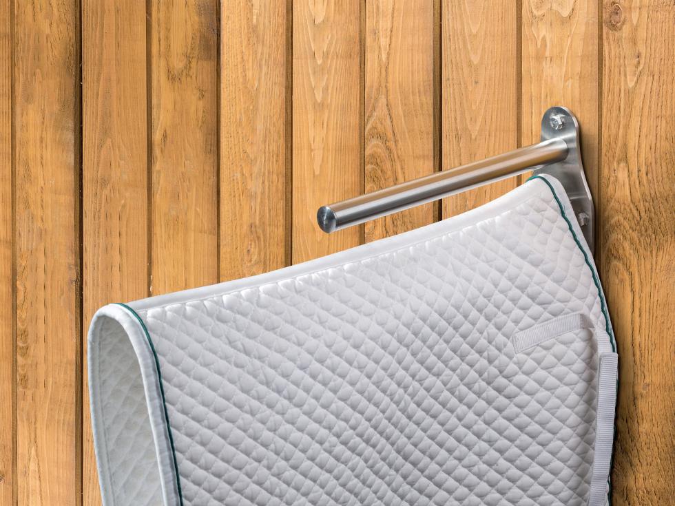 Ein glockenförmiges Flacheisen aus Edelstahl mit solidem Stab als Sattelauflage und darunter mit zwei in umgekehrter V-Form befestigter, länglichen Bügeln zur Auflage einer Schabracke, montiert an einer Holzwand