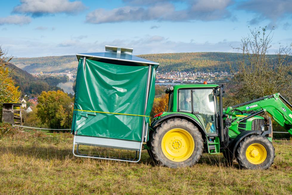 Viereckige verzinkte Futterraufe mit auf dem Grundgerüst befestigten Fressgittern und achteckige Dachkonstruktion aus grüner Plane, wird mit einem Rundballen befüllt durch grünen Traktor mit Frontlader, Weide, Bäume und Himmel im Hintergrund