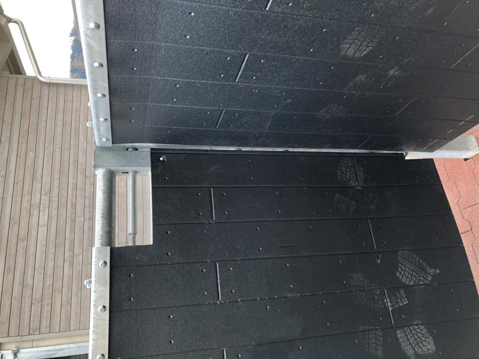 Eine Schlagschutzmatte aus Gummi mit einer Holzbohlenoptik, auf einer Leiste an einer Holzwand montiert, auf der rechten Seite eine Boxenabtrennung einer Pferdebox mit montierter Tränke