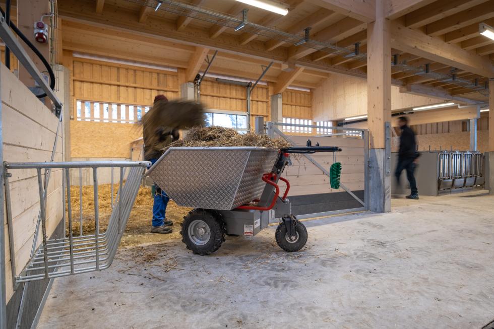 Eine Schubkarre mit Metallmulde, rot lackiertem Rohrgestänge, daraus zwei schwarz gummierte Lenkgriffe, metallener Motorkasten unterlegt, zwei festen Rädern vorne, mit Ackerstollen bereift, einem schwenkbaren Rad hinten, auf einer Baustelle, eine Person an den Griffen, wird von einem Bagger befüllt