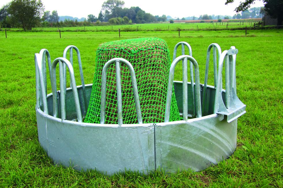 Eine Detailansicht einer runden Rundbogengitterraufe auf Gras, grünes Futtersparnetz überzieht teilweise sichtbarer Futterhaufen und ist an Spannrahmen befestigt, welcher auf dem Boden liegt, Ansicht von oben, zeigt die Hälfte des Innern der Raufe 