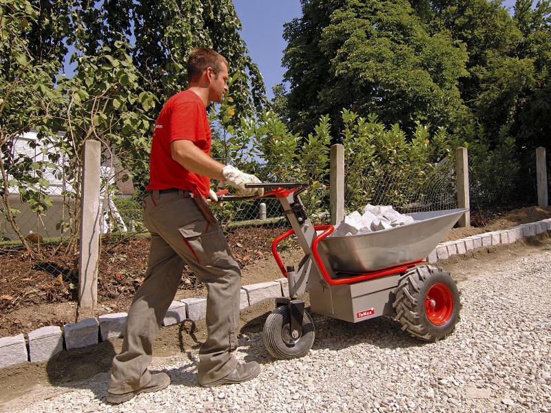 Eine Schubkarre mit Metallmulde, rot lackiertem Rohrgestänge, daraus zwei schwarz gummierte Lenkgriffe, metallener Motorkasten unterlegt, zwei festen Rädern vorne, mit Ackerstollen bereift, einem schwenkbaren Rad hinten, auf einer Baustelle, eine Person an den Griffen, wird von einem Bagger befüllt