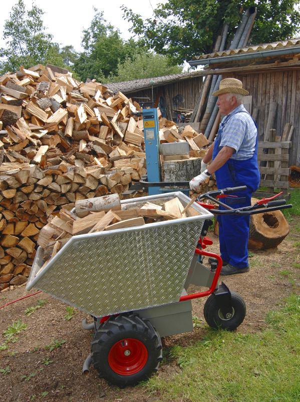 Eine Schubkarre mit Metallmulde, rot lackiertem Rohrgestänge, daraus zwei schwarz gummierte Lenkgriffe, metallener Motorkasten unterlegt, zwei festen Rädern vorne, mit Ackerstollen bereift, einem schwenkbaren Rad hinten, auf einer Baustelle, eine Person an den Griffen, wird von einem Bagger befüllt