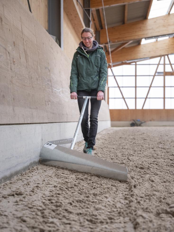 Ein metallischer Hufschlagboy mit einer abgeschrägten Schaufel, einem Metallstiel und einem rechtwinklig darauf montierten Griff, jeweils am Ende schwarz gummiert, wird von einer Person der Kante einer Reithalle nach, durch den Sand gezogen