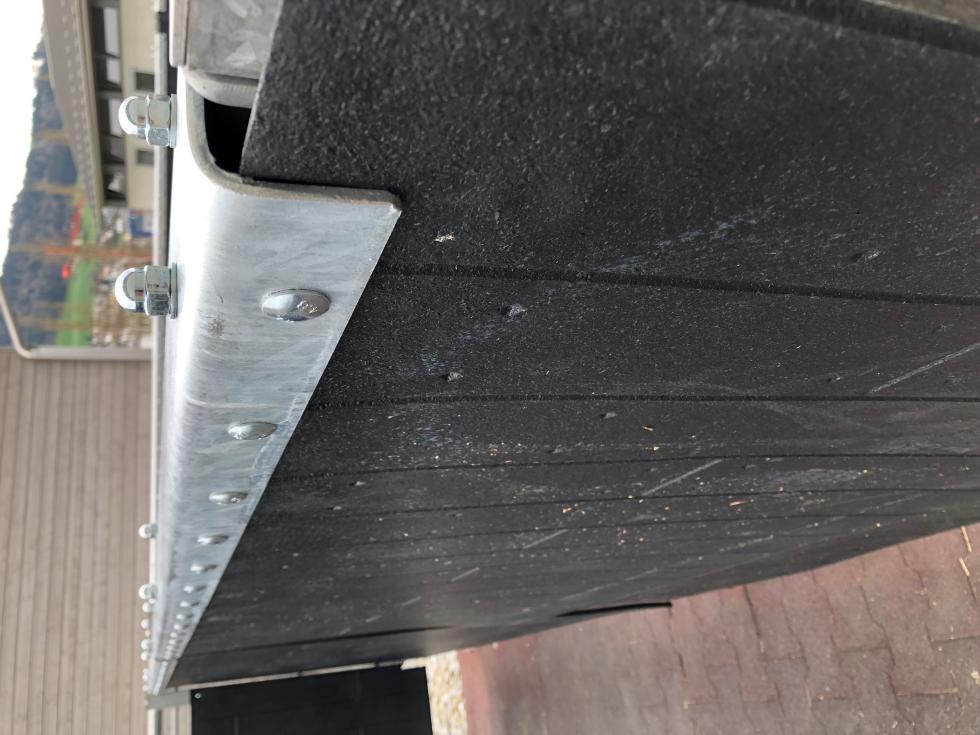 Eine Schlagschutzmatte aus Gummi mit einer Holzbohlenoptik, auf einer Leiste an einer Holzwand montiert, auf der rechten Seite eine Boxenabtrennung einer Pferdebox mit montierter Tränke