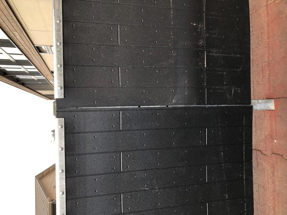 Eine Schlagschutzmatte aus Gummi mit einer Holzbohlenoptik, auf einer Leiste an einer Holzwand montiert, auf der rechten Seite eine Boxenabtrennung einer Pferdebox mit montierter Tränke